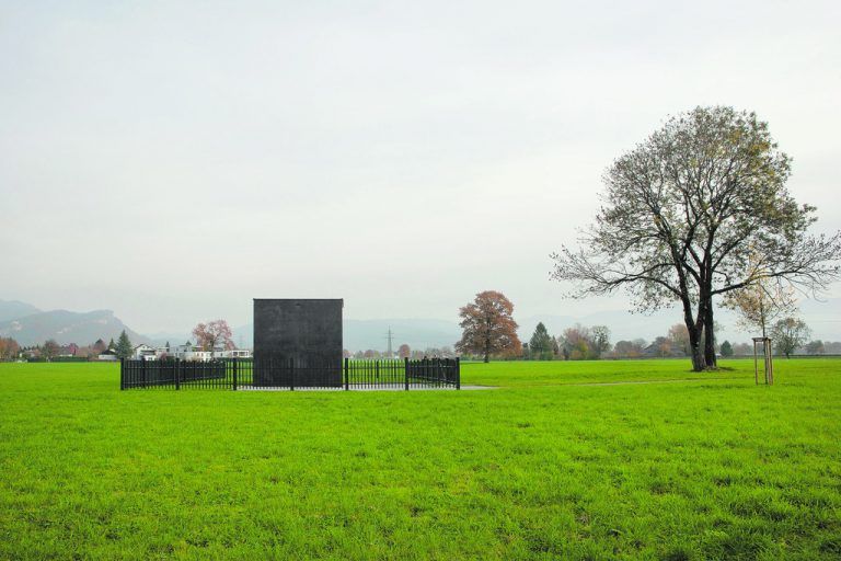 Der Brunnen steht nun als Würfel mit 4,80 Meter Seitenlänge auf einem asphaltierten Platz, der von einem Lattenzaun eingefasst ist.