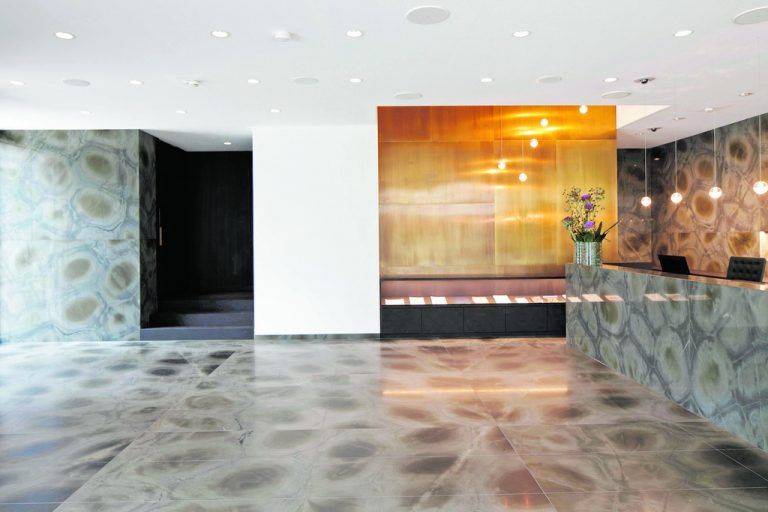 Auch im Foyer ist der Naturstein mit seinem grafischen Muster eindrücklich präsent. Die Messingwand unterstützt die vertikale Wirkung eines neu eingefügten Luftraums. Ein Leuchtenensemble mit perfekt abgestimmter Lichtfarbe trägt zur Atmosphäre dieser Räume wesentlich bei.