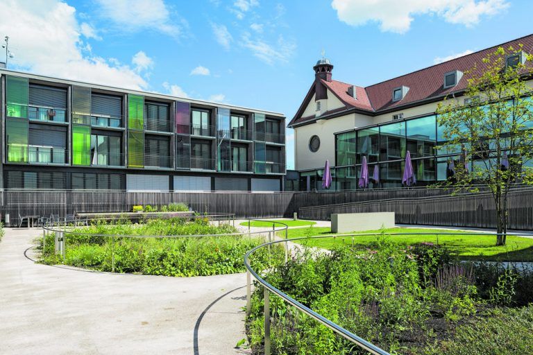 Der Neubau des Pflegeheims schließt das Ensemble zu einem Hof. Die ovalrunden Beete und ein sehr abstrakter Brunnen gliedern eine helle Asphaltfläche.