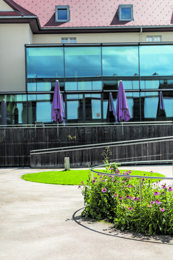 Die spiegelnde Verglasung des Cafés kontrastiert mit der massiven Putzfassade des Bestandsbaus. Die violette Farbgebung setzt sich in den Sitzmöbeln im Inneren fort.