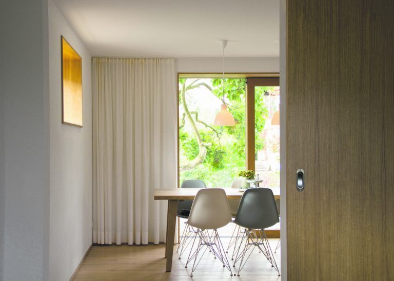 Eichenholzgerahmter Blick auf Terrasse, Baum und Garten. Die Übergänge von innen nach außen, von privatem zu öffentlichem Raum, sind fein abgestuft.