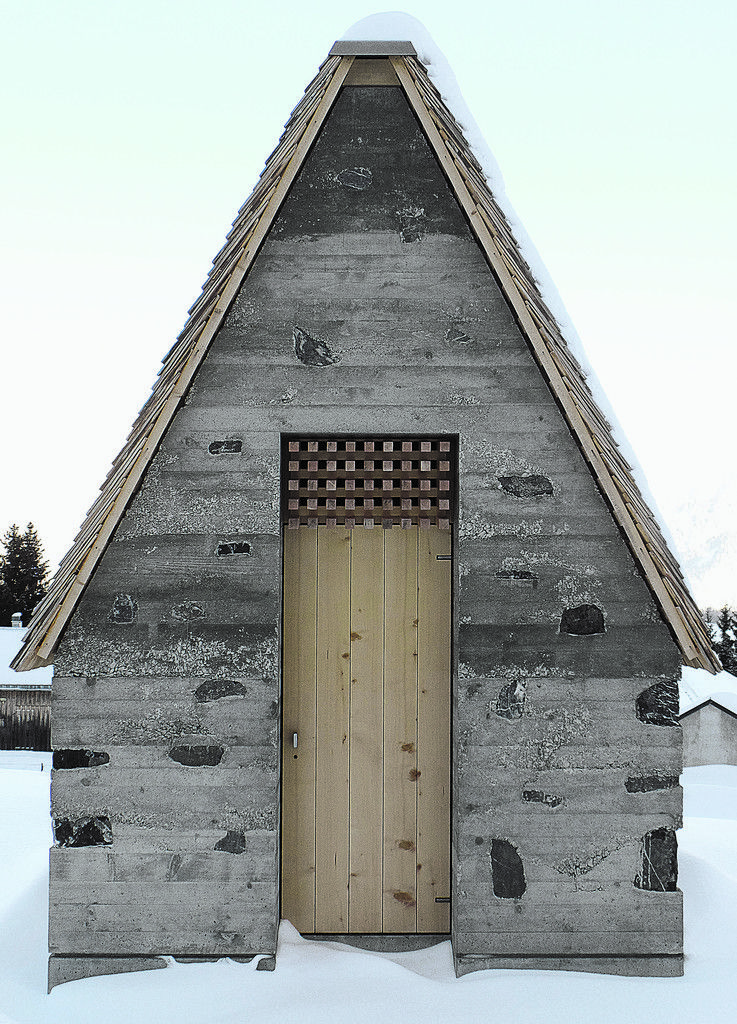 Ein 68° steiles Dach Der First gekappt, um Platz zu machen für die Raumbelichtung; Tür mit Klangraum für die Glocke; Findlinge in der Stampfbetonwand.