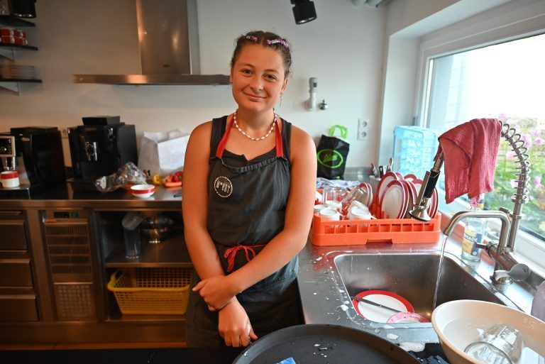Mit Schürze, Charme und einer großen Portion Freude: Emely räumt das Geschirr weg und spült ab.