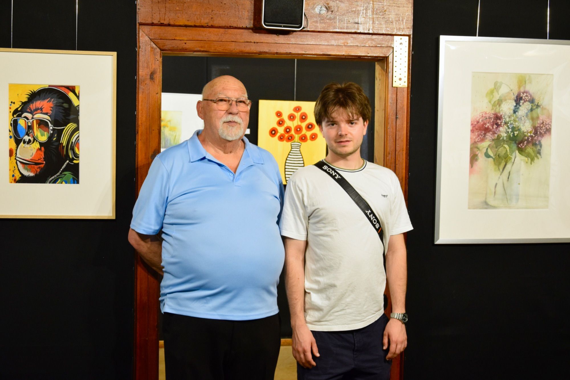 „Haltestelle Kunst“-Initiator 2024 - 2026, Gerhard Montibeller, und David Kathrein („Regio Im Walgau“) zwischen Arbeiten von Sameeh Zaky aus Nenzing („Cicco“, Acryl) und Margot Gabriel aus Frastanz („Flieder“, Aquarell).