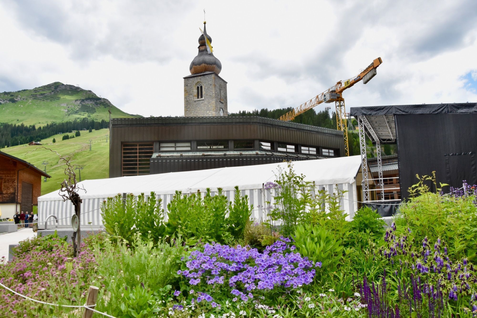 Pflanzen, ein Festzelt, die Neue Pfarrkirche und die jahrhundertealte Walserkirche, umgeben von Bergen.